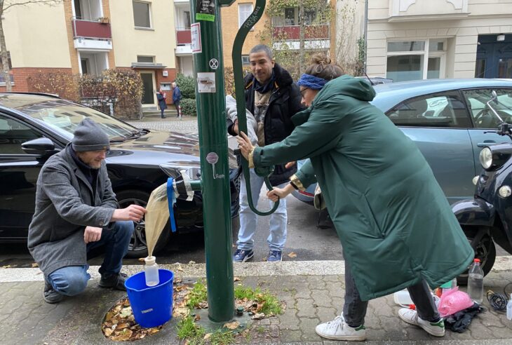 Drei Menschen an einer Schwengelpumpe im Berliner Straßenland. Sie pumpen Grundwasser in ein Filternetz, das am Hahn der Pumpe hängt.
