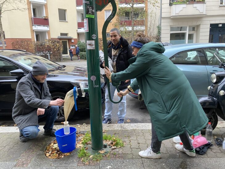 Drei Menschen an einer Schwengelpumpe im Berliner Straßenland. Sie pumpen Grundwasser in ein Filternetz, das am Hahn der Pumpe hängt.
